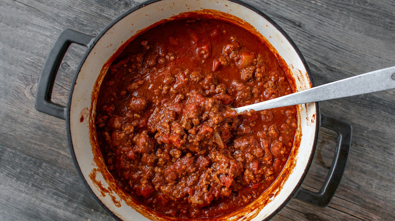 A Dutch oven of chili with a metal serving spoon sticking in it, sitting on a grey wooden surface
