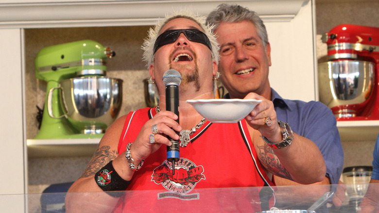 Guy Fieri holds a bowl and a microphone as Anthony Bourdain stands behind him smiling, against a backdrop of several stand-mixers