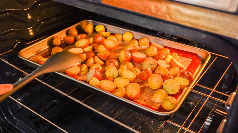 A person uses a long spoon to flip over potatoes that are roasting in the oven on a sheet pan
