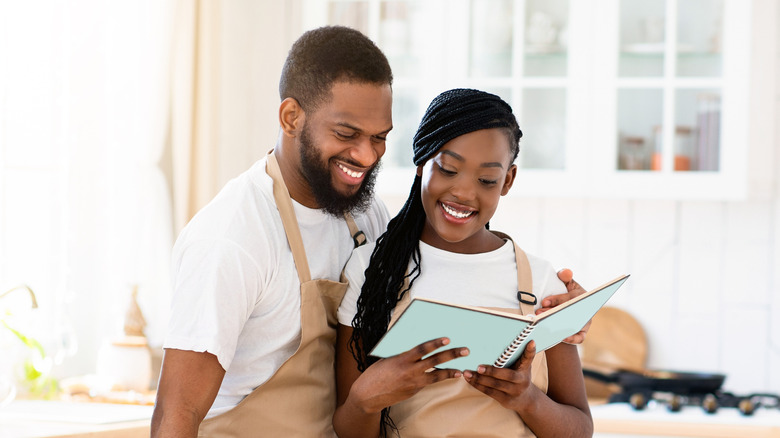 Black couple in a kitchen smiling wearing aprons and looking at a blue notebook
