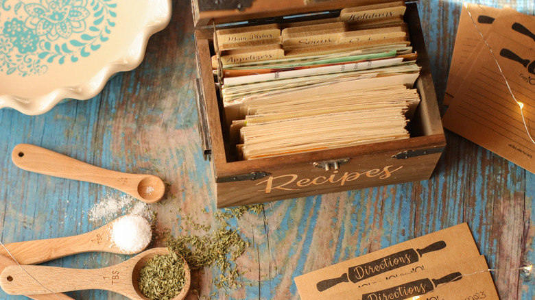 A wood keepsake box of old recipe notecards on a rustic countertop next to wooden spoons with spices