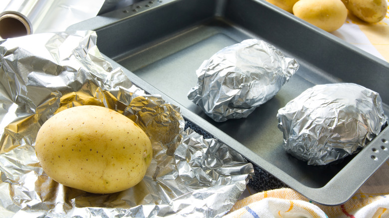 Potatoes in foil wrapped in a baking pan next to an unwrapped potato sitting on foil
