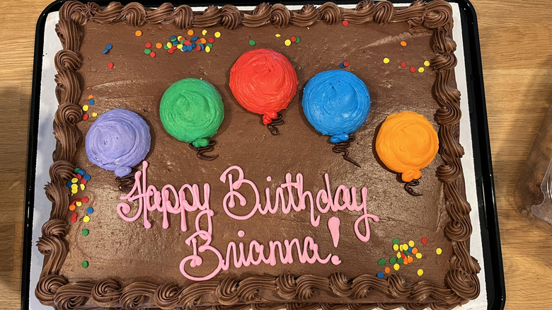 A sheet cake covered in chocolate frosting and some sprinkles with Happy Birthday Brianna written in pink icing, and featuring five colorful icing balloons