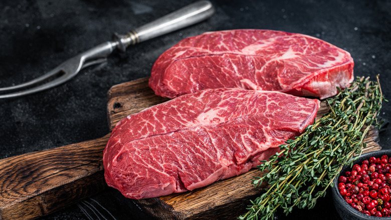 Raw flat iron steaks on wooden board next to thyme sprigs, red peppercorns, and utensil