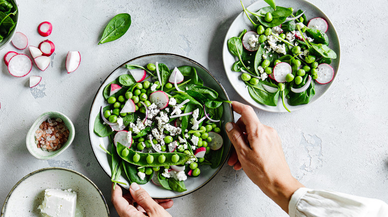 Hands holding white bowl full of snap pea salad next to smaller bowl of salad on white surface