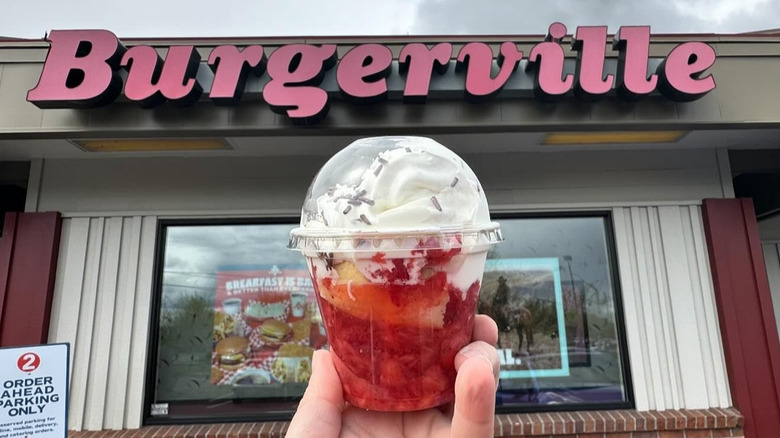 Someone holding a strawberry shortcake in a cup in front of a Burgerville location