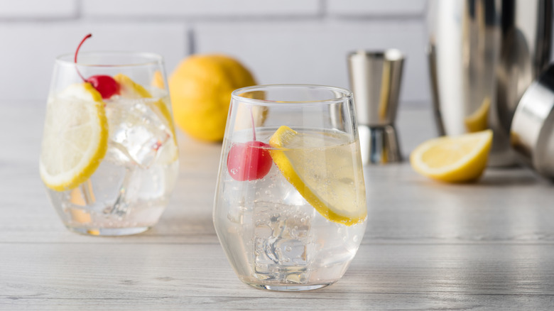 two Tom Collins cocktails with lemons and cherries and a cocktail shaker in the background