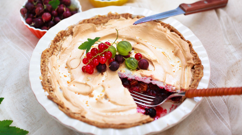 A white pie pan is filled with a golden brown crust and a whipped topping topped with gooseberries. A slice is missing to reveal the flavor of the pie is the same gooseberries that are on top of the pie,