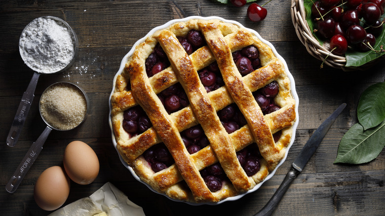 A dark brown wooden table has ingredients including two eggs, butter, a measuring cup filled with sugar, flour, and a bowl full of cherries, surrounding a cherry pie with a lattice crust