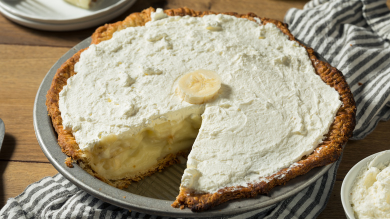 A silver pie tin sits on a light brown wooden table and a blue and white striped towel. The pie tin is filled with a brown buttery crust, a custardy yellow banana filling, whipped cream, and a sliced banana