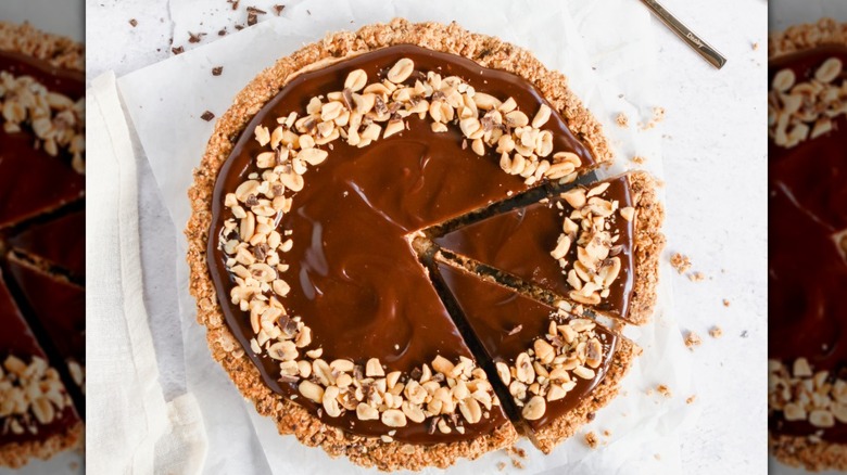 A white marble countertop and white towel are topped with a chocolate peanut butter pie that is topped with additional peanuts to reflect the creamy peanut butter filling. Two slices are cut and slightly pulled back to show off the peanut butter filling.