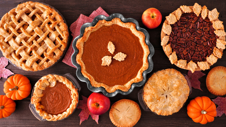 A dark brown wooden table is adorned with mini decorative orange pumpkins, red apples, and red maple leaves, along with four mini pies and three larger pies each of a different pie filling and topped with a different pie crust style to reflect the diversity of pie offerings in the fall