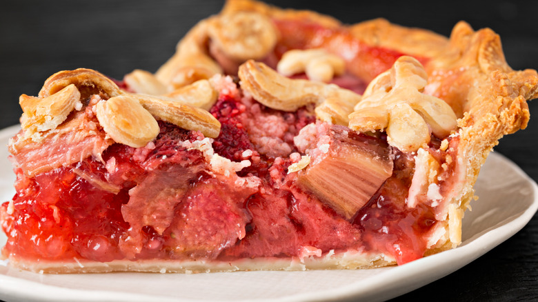 A closeup of a single slice of pie on a white plate details the buttery golden brown crust, strawberries, and rhubarb filling in a vibrant shade of pink