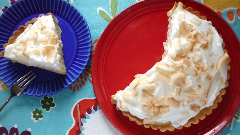 A blue plate is topped with a silver fork and a single slice of pie next to a larger red plate topped with the remaining larger pie. The pie is white, fluffy, whipped cream looking in texture and topped with shaved toasted coconut