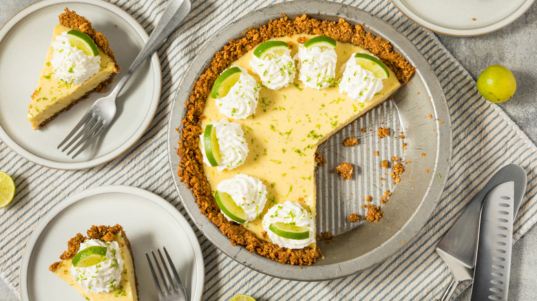 A silver pie tine with a vibrant yellow-green filling, topped with dollops of whipped cream and small slices of green limes has a few slices missing. Next to the pie pan are three white plates topped with a silver fork and a single slice of the pie from the larger pie pan.
