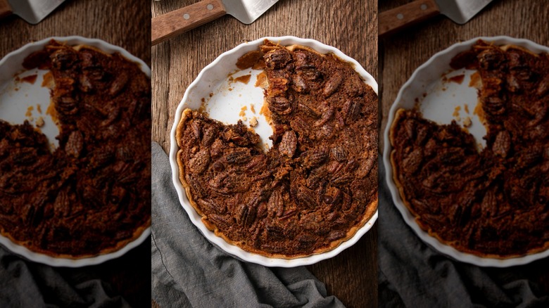 A white pie pan is filled with a dark chocolate pecan pie mixture with a slice missing to reveal the flaky texture of the pie crust