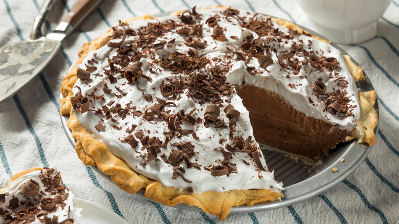 A white towel with blue stripes is topped with a French silk pie with a single slice cut out to visually show off the fluffy chocolate mixture, the buttery crust, and the whipped topping covered with chocolate shavings