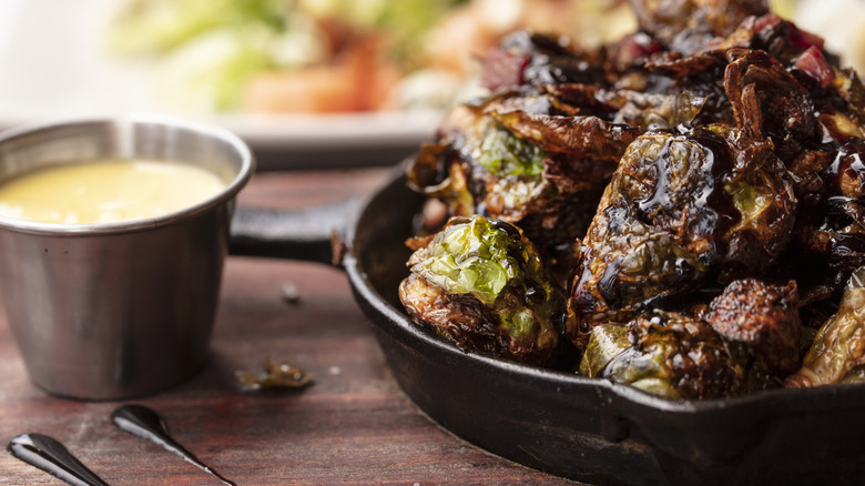 A cast iron pan of blackened Brussels sprouts sits on a wooden board with dipping sauce next to it.