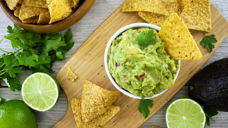 Bowl of guacamole and chips on board next to avocados, limes, and cilantro