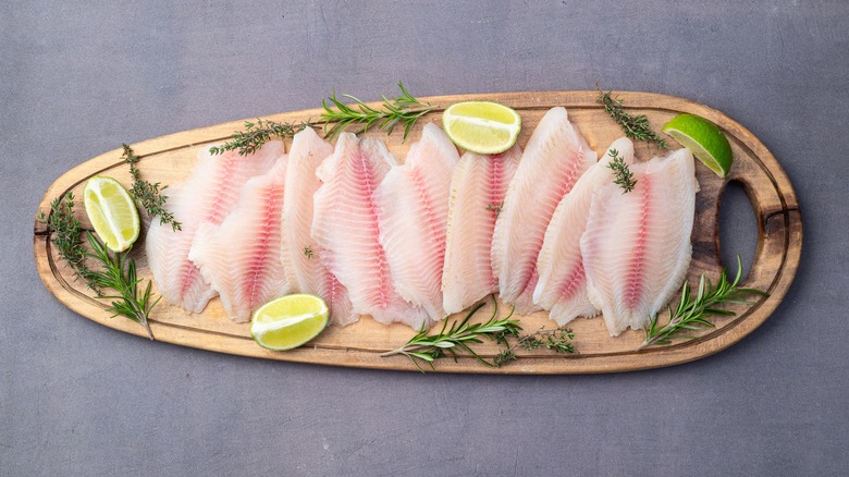 Tilapia filets, raw, on a cutting board garnished with herbs and lime
