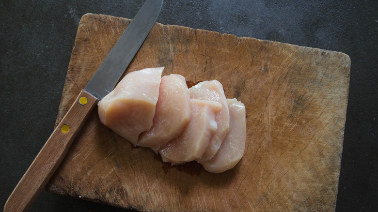 Sliced raw chicken breast on wooden cutting board with knife