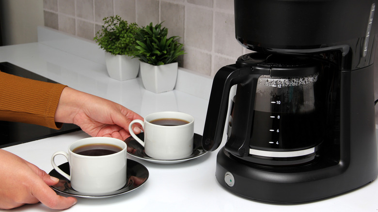 An automatic drip coffee machine in black on a white table with two coffee cups