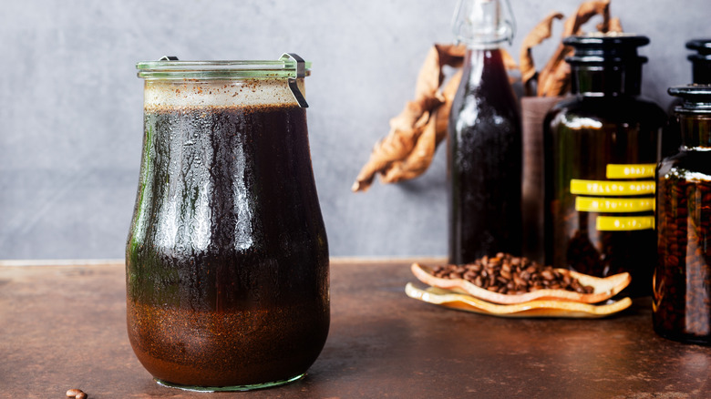 A jar of coffee grounds steeping in water on a table with other jars and more coffee beans