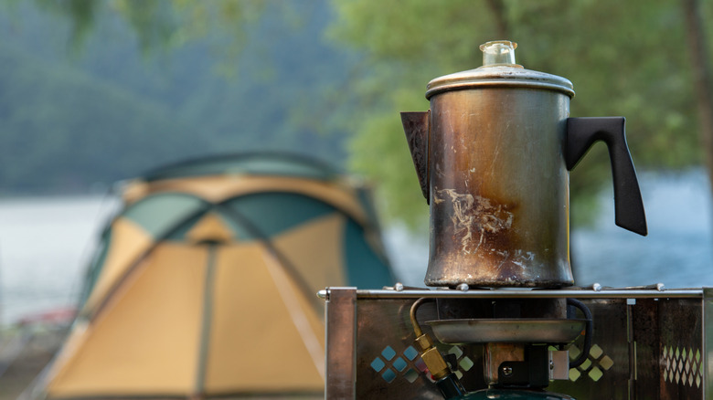 A stainless steel percolator on a camp stove outside with a tent in the background