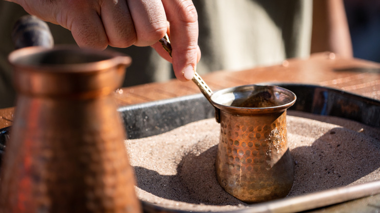 A close-up of a hand holding the handle of a cezve sitting in hot sand