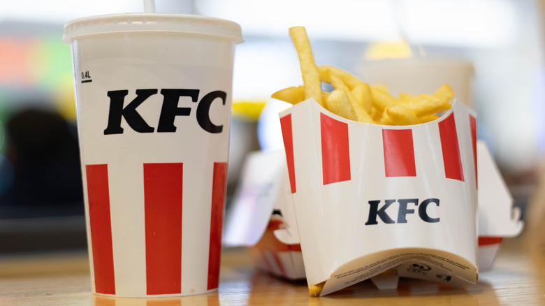 a KFC-branded fountain drink and cardboard container of french fries on a wooden table with a box and bag in the background