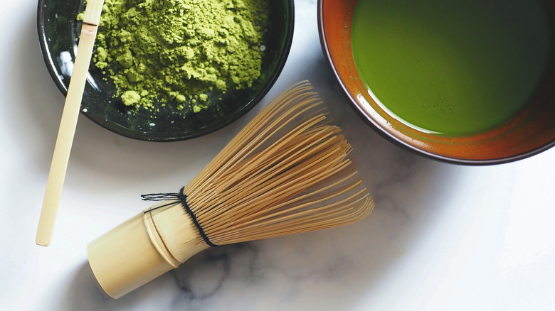A bamboo matcha whisk with a bowl of matcha powder alongside another bowl with matcha powder that's been whisked with water all on a marble countertop