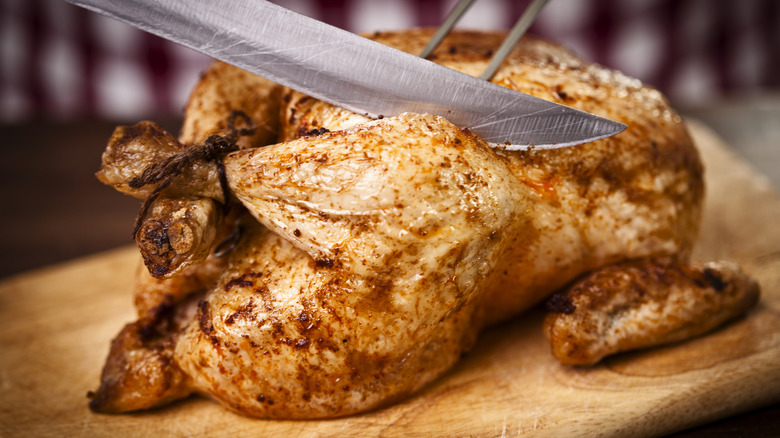 a roast chicken on a wooden cutting board being carved with a carving fork and a long knife