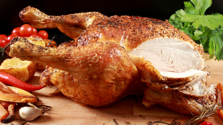 a roast chicken with the breast sliced open revealing the white meat beneath on a cutting board with herbs and vegetables