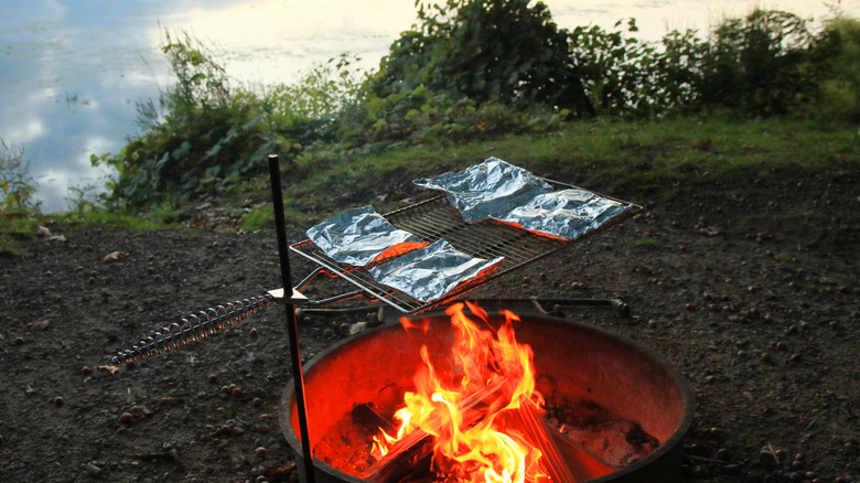 Four foil packet meals cooking over a campfire by a lakeside