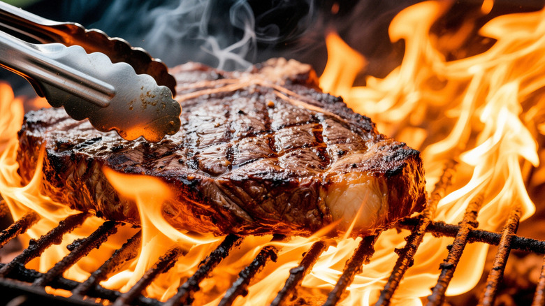 A T-bone steak on a grill among flames, metal tongs pressing it down