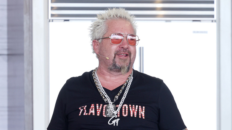 "Diners, Drive-Ins and Dives" host Guy Fieri stands and smiles while wearing a black t-shirt reading "Flavortown"