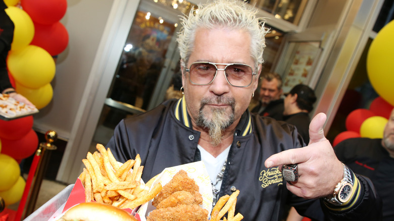 "Diners, Drive-Ins and Dives" host Guy Fieri holds a platter containing a sandwich, fried chicken tenders, and French fries while pointing at it with his other hand