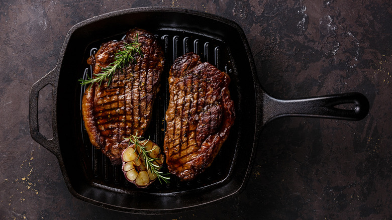 Two large grilled steaks in a cast iron pan alongside rosemary and garlic