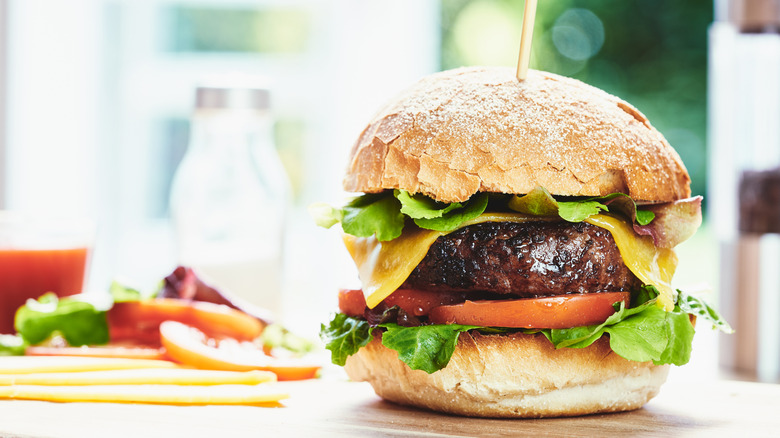 A grilled cheeseburger on a stacked bun with ingredients seen in the background