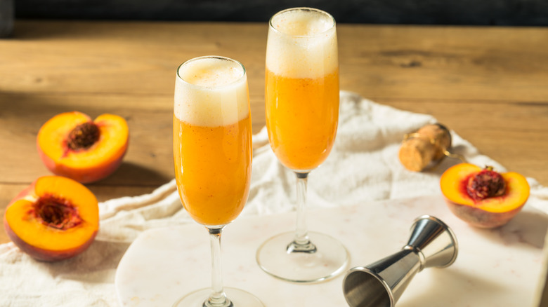 Two peach bellinis alongside two fresh peaches on a light colored cloth and a white background