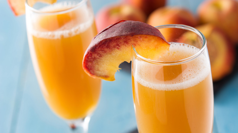 Two bellinis with a jigger, halved peaches, and a wine cork all on top of a white cloth on a wooden table