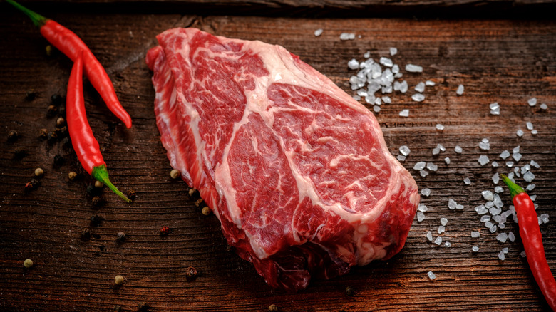 raw beef chuck roast on a wooden cutting board with coarse salt, peppercorns, and red peppers.