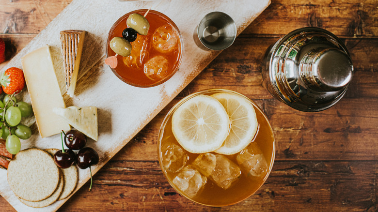 two different cocktails and a shaker on a wood table with cheese and fruit
