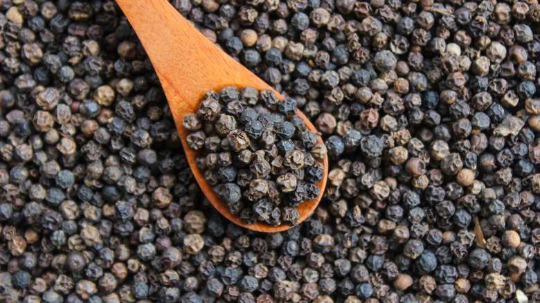 Wooden spoon full of black peppercorns atop pile of peppercorns.