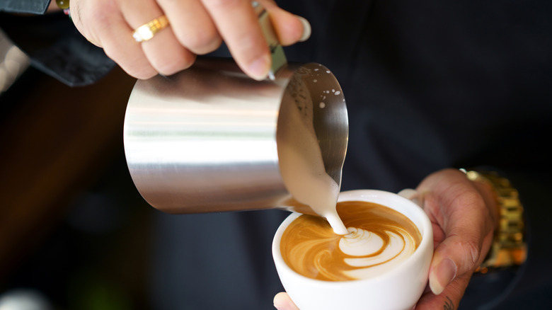 Barista hands pouring milk foam into espresso resulting in design of latte