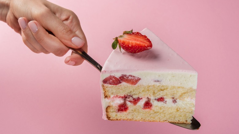 Hand holding a slice of strawberry cake on a spatula with a pink background
