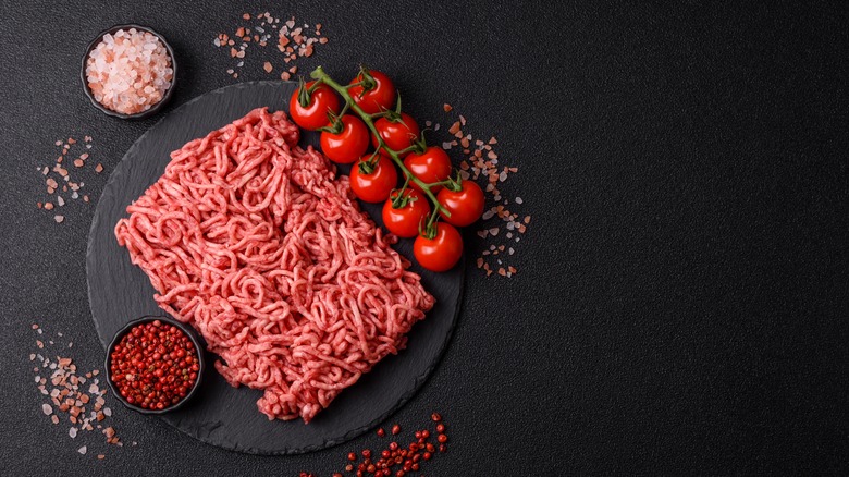Raw ground beef with salt, peppercorns, and tomatoes surrounding it on a black table
