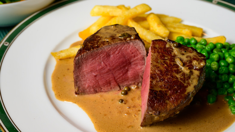 Medium rare filet mignon steak cut in half and served over sauce with french fries and peas