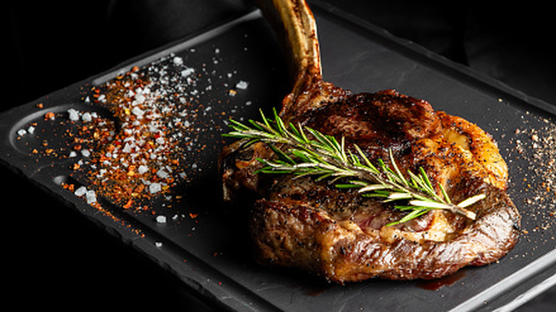 overhead view of a riebeye topped with salt, pepper, and a sprig of fresh rosemary on a white plate with a fork and knife beside it