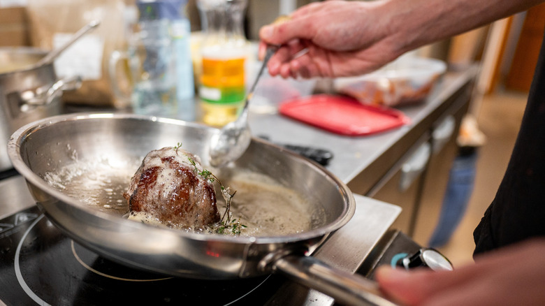 a cook bastes a steak with butter in a stainless steel pan